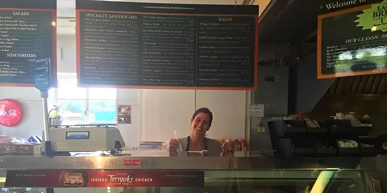 Smiling woman behind sandwich shop counter.